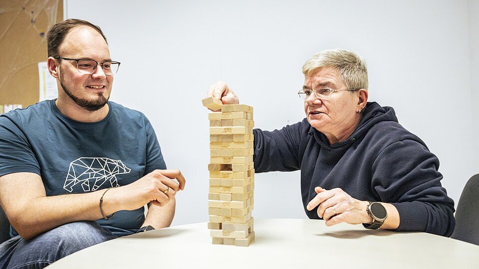 Zwei Personen unterschiedlichen Alters spielen gemeinsam Jenga an einem Tisch und konzentrieren sich auf den wackelnden Holzturm.