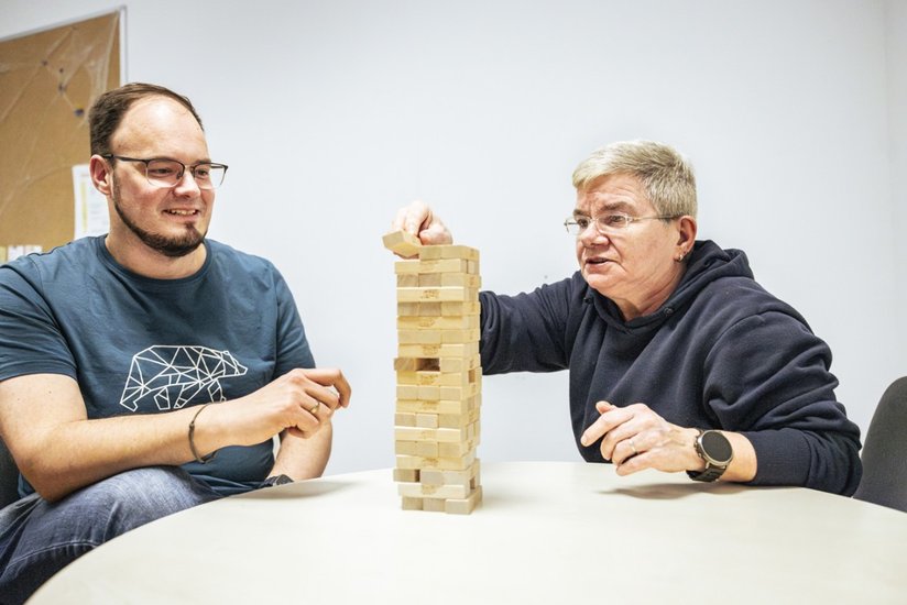 Zwei Personen unterschiedlichen Alters spielen gemeinsam Jenga an einem Tisch und konzentrieren sich auf den wackelnden Holzturm.