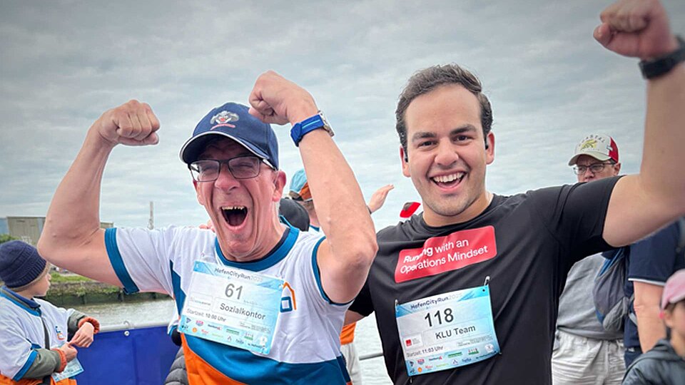 Zwei Männer feiern nach dem HafenCity Run mit erhobenen Armen und breitem Lächeln. Der eine trägt ein Sozialkontor-Shirt mit Startnummer 61, der andere ein schwarzes Klu-Team-Shirt mit der Startnummer 118 und der Aufschrift „Running with an Operations Mindset“.
