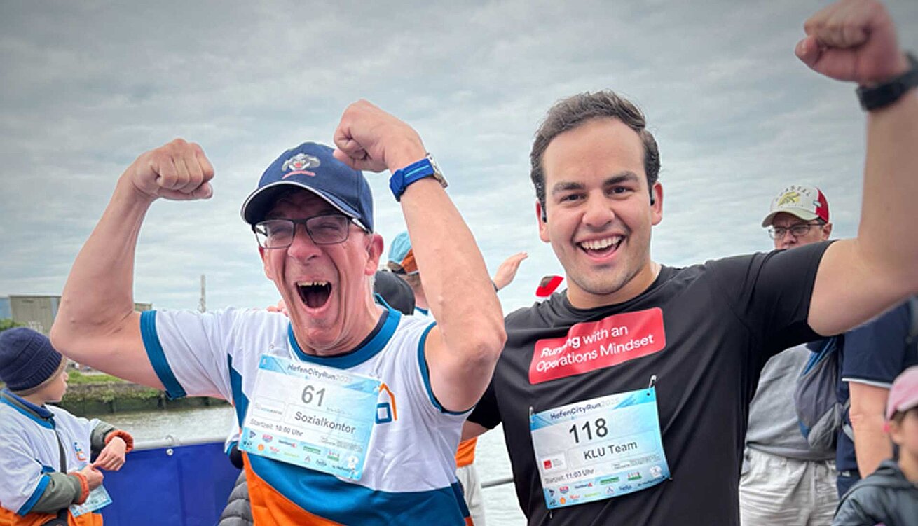 Zwei Männer feiern nach dem HafenCity Run mit erhobenen Armen und breitem Lächeln. Der eine trägt ein Sozialkontor-Shirt mit Startnummer 61, der andere ein schwarzes Klu-Team-Shirt mit der Startnummer 118 und der Aufschrift „Running with an Operations Mindset“.