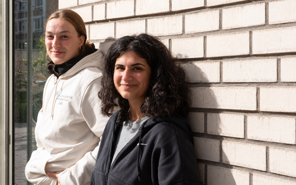 Was uns ausmacht im Sozialkontor: Zwei lächelnde Frauen stehen nebeneinander vor einer hellen Backsteinwand. Eine trägt einen weißen Hoodie mit Sozialkontor-Logo.