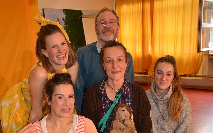 Fünf Personen posieren für ein Foto in einem Raum mit Vorhängen, in dem eine Frau einen Stoffigel in der Hand hält.