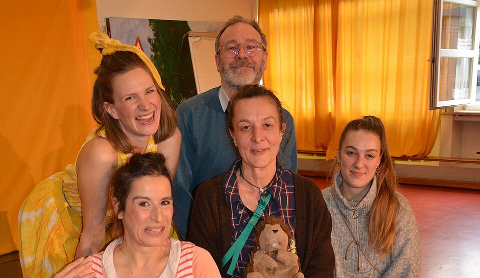 Fünf Personen posieren für ein Foto in einem Raum mit Vorhängen, in dem eine Frau einen Stoffigel in der Hand hält.