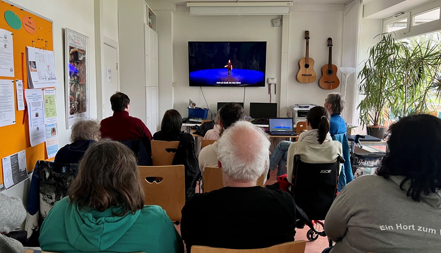 Eine Gruppe von Menschen die sich auf einem großen Bildschirm eine Vorstellung von Theater ohne Grenzen anguckt