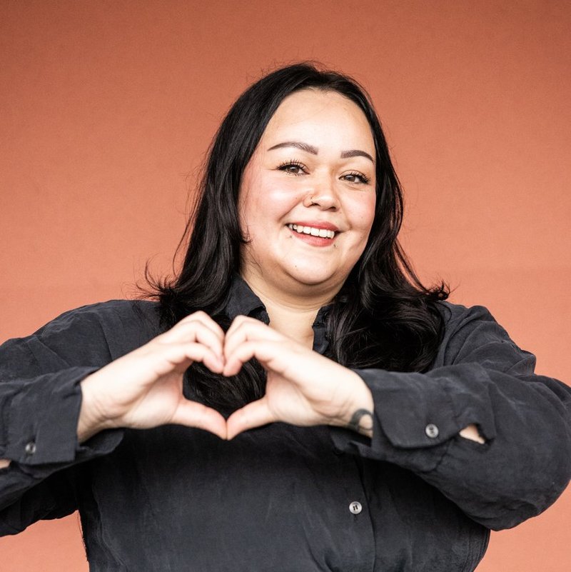 Eine Frau mit dunklen langen Haaren lächelt und formt mit beiden Händen ein Herz vor ihrem Brustkorb. Sie steht vor einem orangefarbenen Hintergrund.