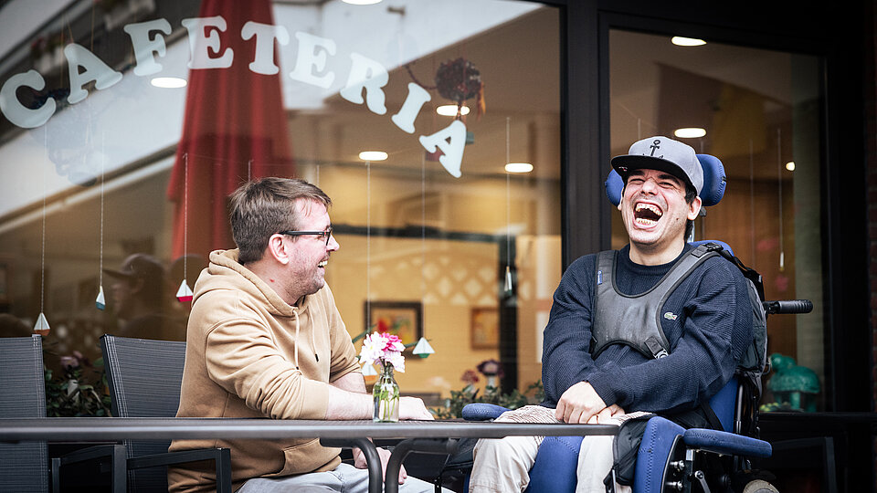 Vor einer Cafeteria sitzen zwei Männer an einem Tisch und lachen gemeinsam. Links sitzt ein Mann mit Brille und beigefarbenem Kapuzenpullover, rechts ein junger Mann im elektrischen Rollstuhl mit dunkelblauem Pulli, hellem Basecap und Armstützen. Beide wirken gelöst und fröhlich. Auf dem Tisch steht eine kleine Blumenvase. Im Hintergrund ist durch das Fenster der Innenbereich der Cafeteria zu sehen.