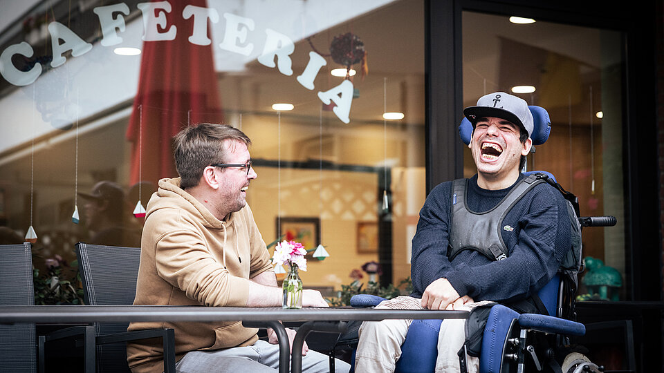 Vor einer Cafeteria sitzen zwei Männer an einem Tisch und lachen gemeinsam. Links sitzt ein Mann mit Brille und beigefarbenem Kapuzenpullover, rechts ein junger Mann im elektrischen Rollstuhl mit dunkelblauem Pulli, hellem Basecap und Armstützen. Beide wirken gelöst und fröhlich. Auf dem Tisch steht eine kleine Blumenvase. Im Hintergrund ist durch das Fenster der Innenbereich der Cafeteria zu sehen.
