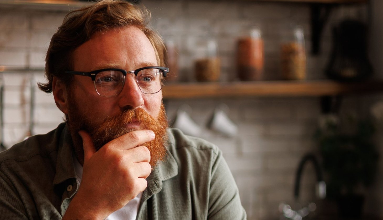 Eine Person mit kurzen roten Haaren, rotem Bart und Brille, fasst sich ans Kinn und schaut nachdenklich in die Ferne. Im Hintergrund ist unscharf eine Küche zu sehen.