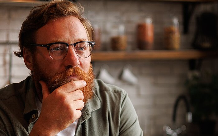Eine Person mit kurzen roten Haaren, rotem Bart und Brille, fasst sich ans Kinn und schaut nachdenklich in die Ferne. Im Hintergrund ist unscharf eine Küche zu sehen.