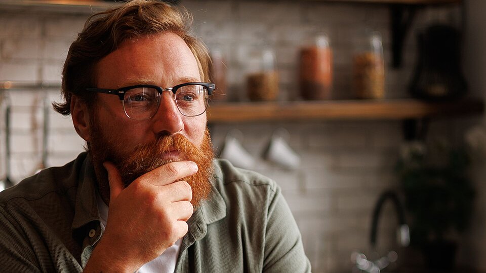 Eine Person mit kurzen roten Haaren, rotem Bart und Brille, fasst sich ans Kinn und schaut nachdenklich in die Ferne. Im Hintergrund ist unscharf eine Küche zu sehen.