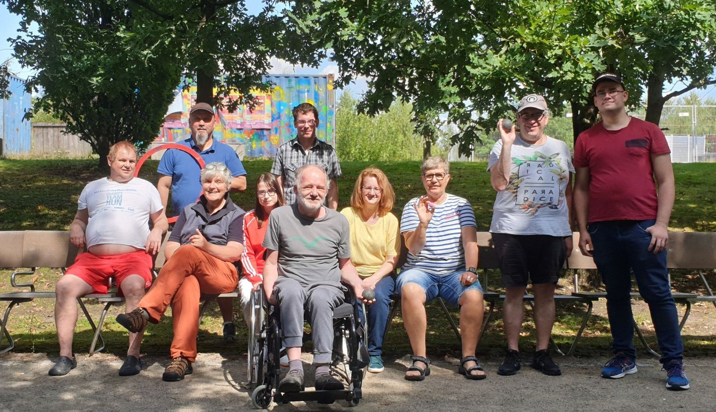 Zehn Erwachsene verschiedenen Alters in sommerlicher Freizeitkleidung. Ein Mann sitzt im Rollstuhl. Das ist die Boule-Gruppe Baaken Bande vom Sozialkontor im Lohsepark in Hamburg.