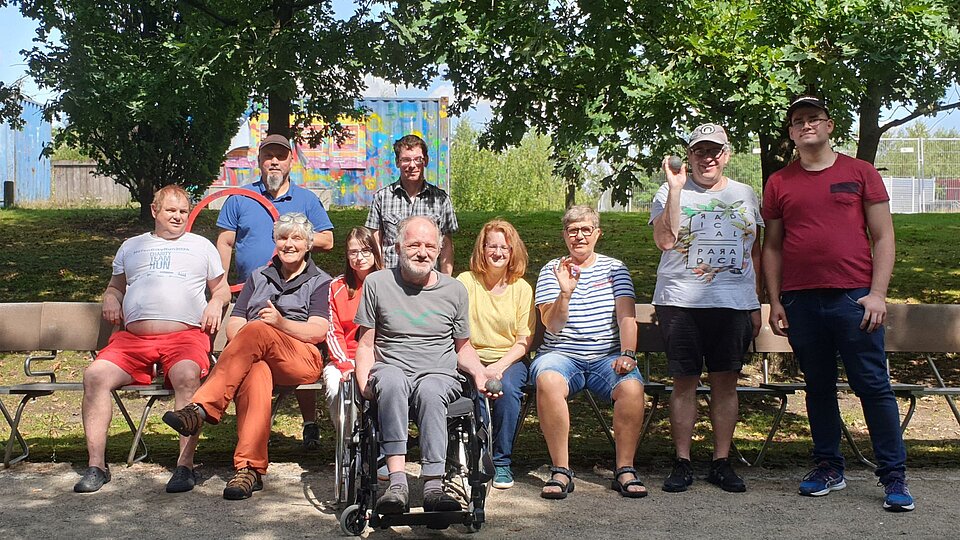 Zehn Erwachsene verschiedenen Alters in sommerlicher Freizeitkleidung. Ein Mann sitzt im Rollstuhl. Das ist die Boule-Gruppe Baaken Bande vom Sozialkontor im Lohsepark in Hamburg.