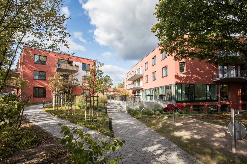 Außenansicht einer modernen, barrierefreien Wohnanlage mit zwei rot verklinkerten Gebäuden, breiten Wegen und viel Grün. Kleine Bäume und Sträucher säumen die Wege, die durch den begrünten Innenhof führen. Die Szene wirkt ruhig und einladend und zeigt ein Wohnangebot des Sozialkontors für Menschen mit Unterstützungsbedarf in einem inklusiven Umfeld.