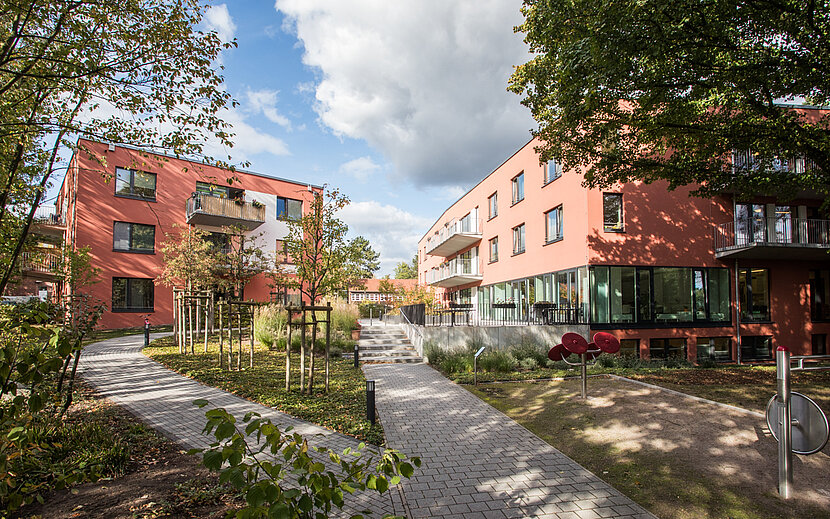 Außenansicht einer modernen, barrierefreien Wohnanlage mit zwei rot verklinkerten Gebäuden, breiten Wegen und viel Grün. Kleine Bäume und Sträucher säumen die Wege, die durch den begrünten Innenhof führen. Die Szene wirkt ruhig und einladend und zeigt ein Wohnangebot des Sozialkontors für Menschen mit Unterstützungsbedarf in einem inklusiven Umfeld.