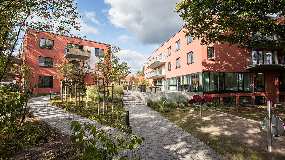 Außenansicht einer modernen, barrierefreien Wohnanlage mit zwei rot verklinkerten Gebäuden, breiten Wegen und viel Grün. Kleine Bäume und Sträucher säumen die Wege, die durch den begrünten Innenhof führen. Die Szene wirkt ruhig und einladend und zeigt ein Wohnangebot des Sozialkontors für Menschen mit Unterstützungsbedarf in einem inklusiven Umfeld.
