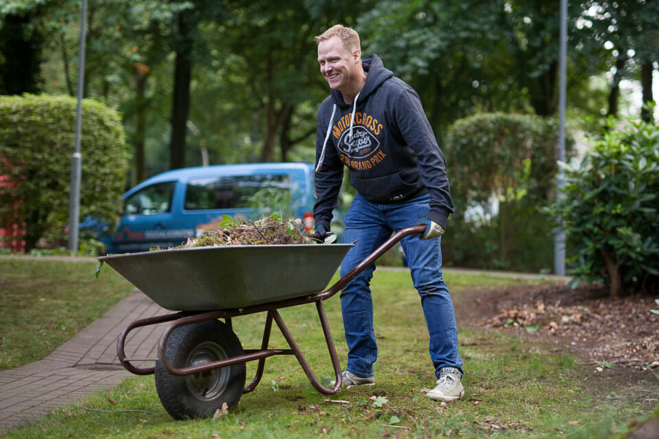 Ein Mann mit einer Schubkarre in der sich Erde und Unkraut befindet.