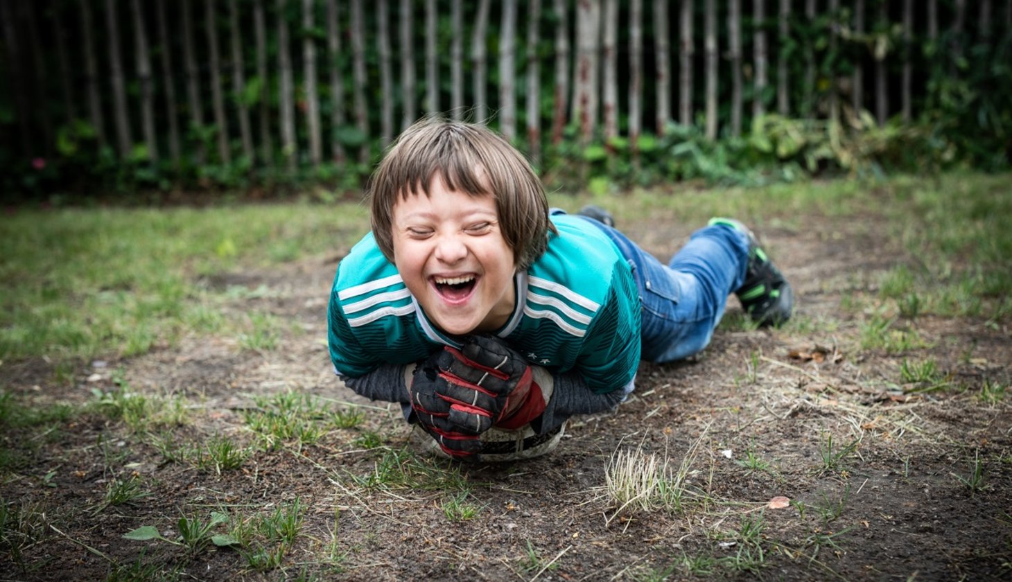 Ein junge liegt lachend auf einen Fußball am Boden auf einem Sportplatz.