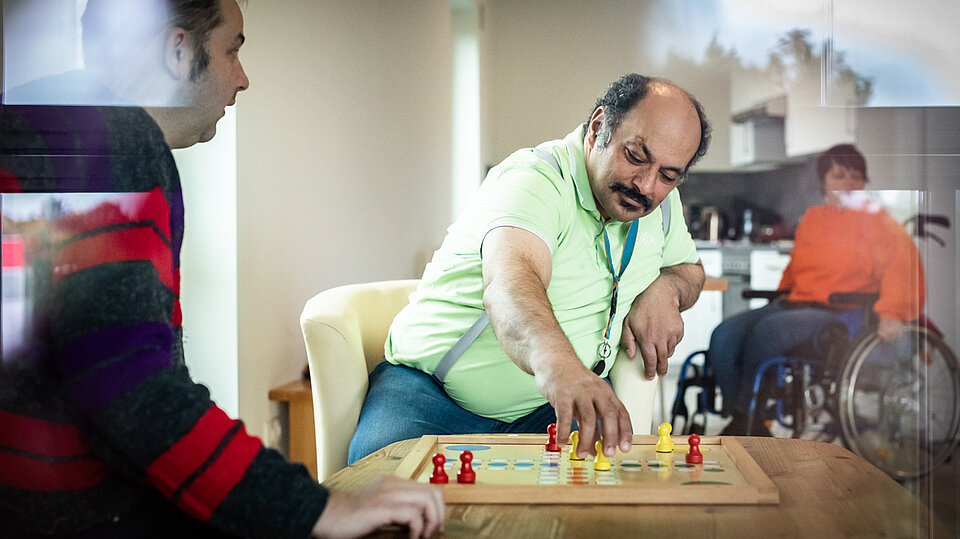 Zwei Männer sitzen an einem Tisch und spielen ein Brettspiel. Einer von ihnen greift konzentriert nach einer Spielfigur. Im Hintergrund ist eine weitere Person im Rollstuhl zu sehen.