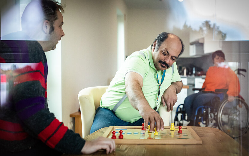 Zwei Männer sitzen an einem Tisch und spielen ein Brettspiel. Einer von ihnen greift konzentriert nach einer Spielfigur. Im Hintergrund ist eine weitere Person im Rollstuhl zu sehen. 
