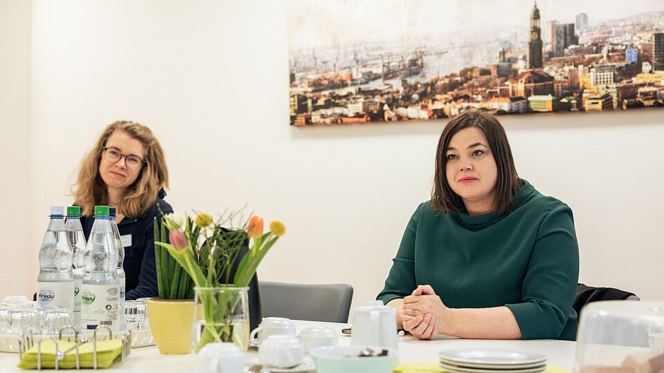 Katharina Fegebank (rechts) und Britt Viemann sitzen an einem Tisch, auf dem Blumen, Wasserflaschen und Geschirr stehen. Im Hintergrund hängt ein Hamburg-Bild an der Wand.