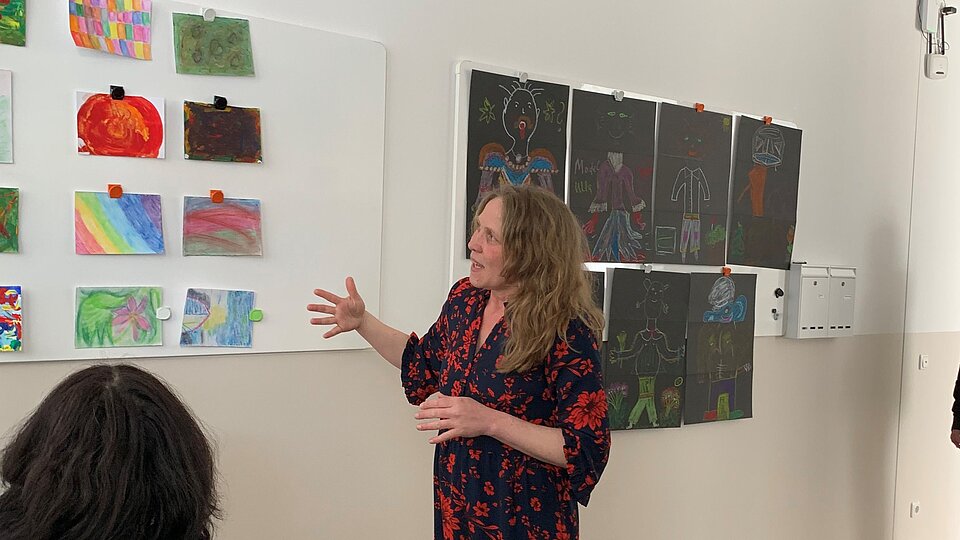 Eine Frau steht vor einer Wand mit Ausstellungen von gemalten Bildern und spricht über diese. Eine andere Frau sitzt im Rollstuhl vor ihr und hört zu.
