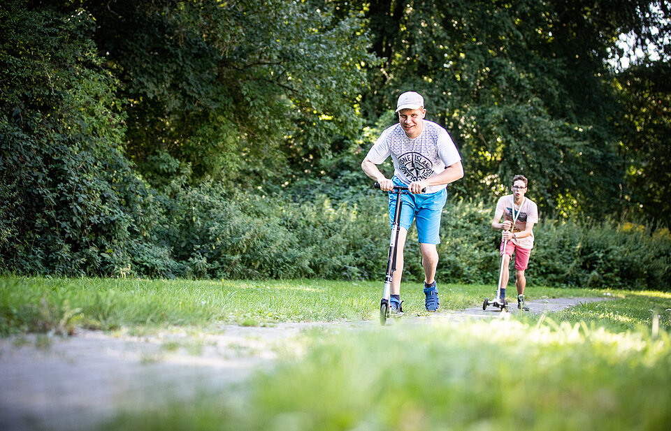 Zwei Jungen machen mit Rollern Sport auf einem Weg durch eine grüne Wiese, umgeben von Bäumen. Der Vordergrund zeigt einen Jungen in blauer Hose und heller Kappe, der aktiv voranfährt.