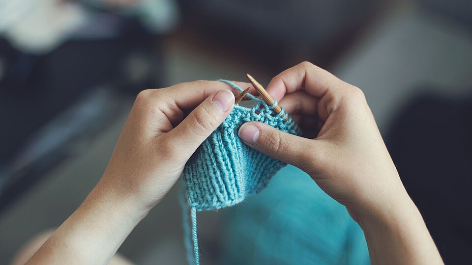 Zwei Hände halten Stricknadeln und stricken mit hellblauer Wolle, im Hintergrund ist ein unscharfer Innenraum zu sehen.