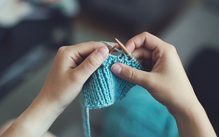 Zwei Hände halten Stricknadeln und stricken mit hellblauer Wolle, im Hintergrund ist ein unscharfer Innenraum zu sehen.