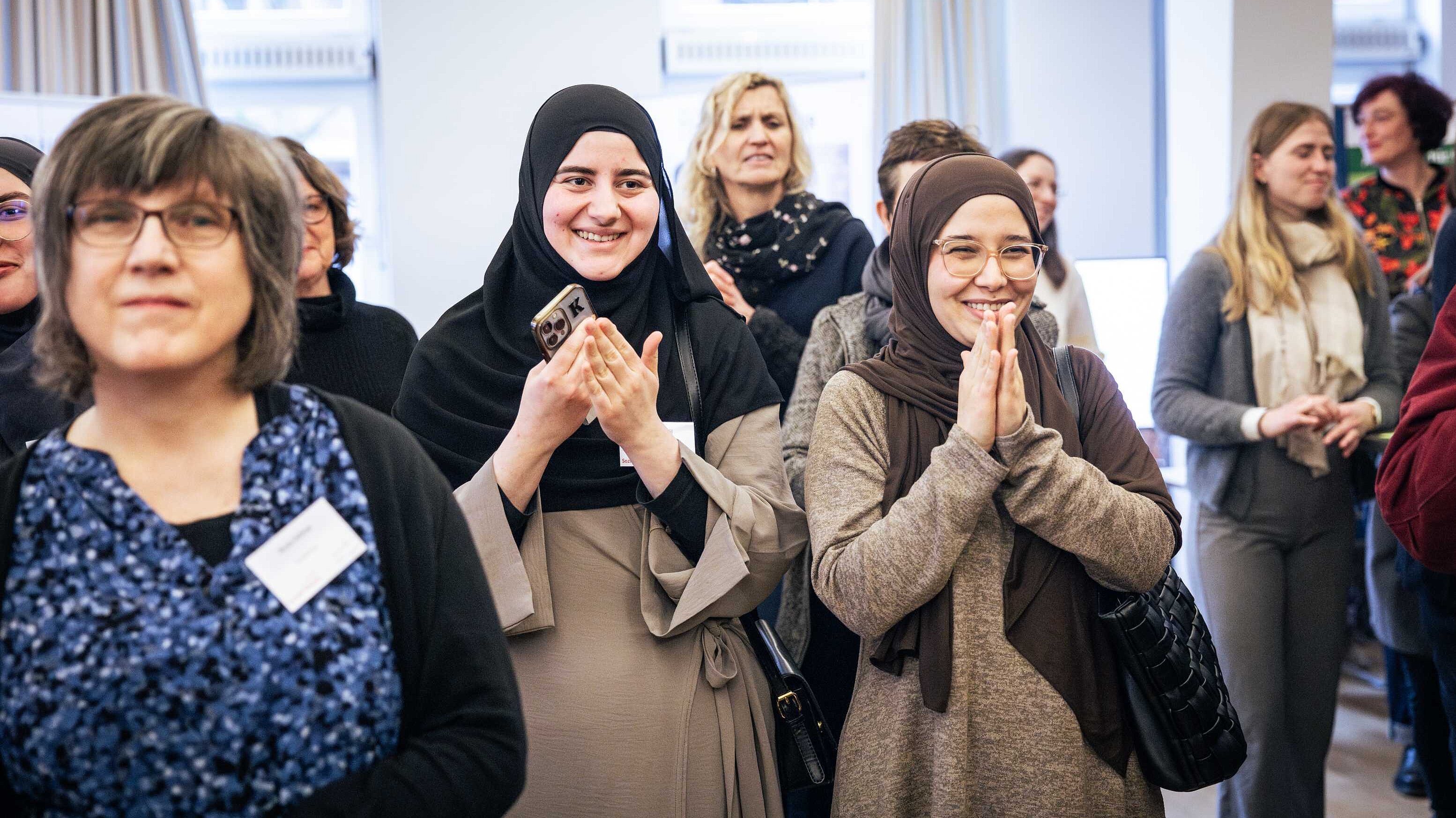 Zwei junge Frauen mit Kopftuch und langen, grauen Kleidern lächeln und applaudieren. Um sie herum sind viele weitere Teilnehmende der Veranstaltung in lebendigem Austausch.