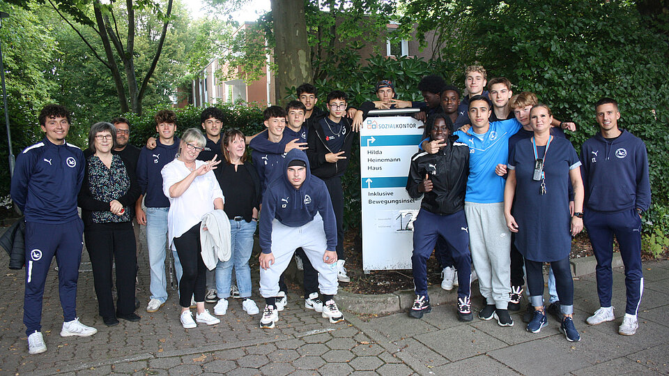 Jugendliche des USC Paloma sowie deren Betreuer*innen und Mitarbeitende des Sozialkontors posieren bei einem Gruppenfoto vor dem Eingangsschild des Hilda Heinemann Haus. Im Hintergrund sind Bäume und ein Haus zu sehen.