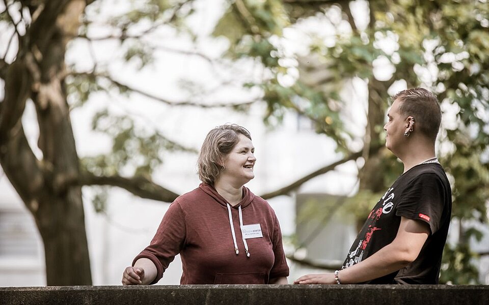 Zwei Personen stehen im Freien an einem Geländer und unterhalten sich angeregt und lachend. Im Hintergrund sind grüne Bäume und ein Gebäude zu sehen.