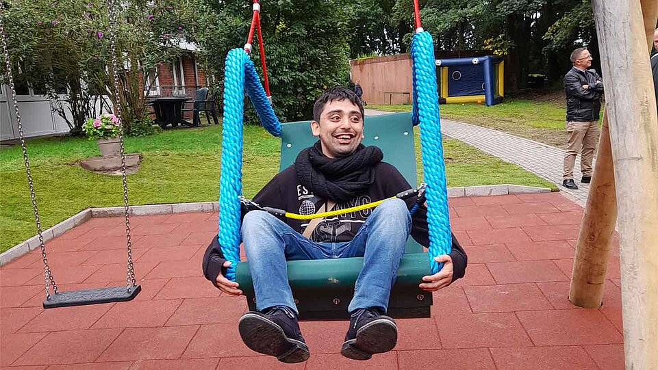 Eine Person sitzt auf einer breiten Schaukel mit blauen Seilen auf einem Spielplatz. Im Hintergrund sind Rasenflächen, Bäume und weitere Spielgeräte zu sehen.