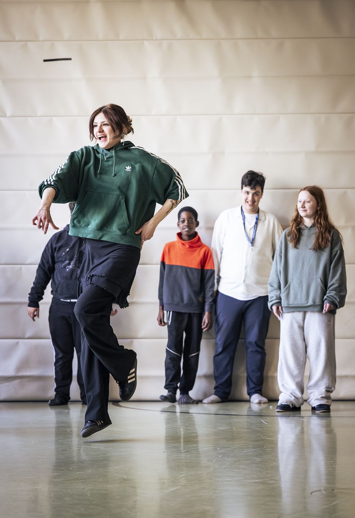 Eine junge Frau in sportlicher Kleidung führt energisch eine Hip-Hop-Tanzbewegung aus. Im Hintergrund stehen mehrere Kinder und Jugendliche und schauen ihr begeistert zu.
