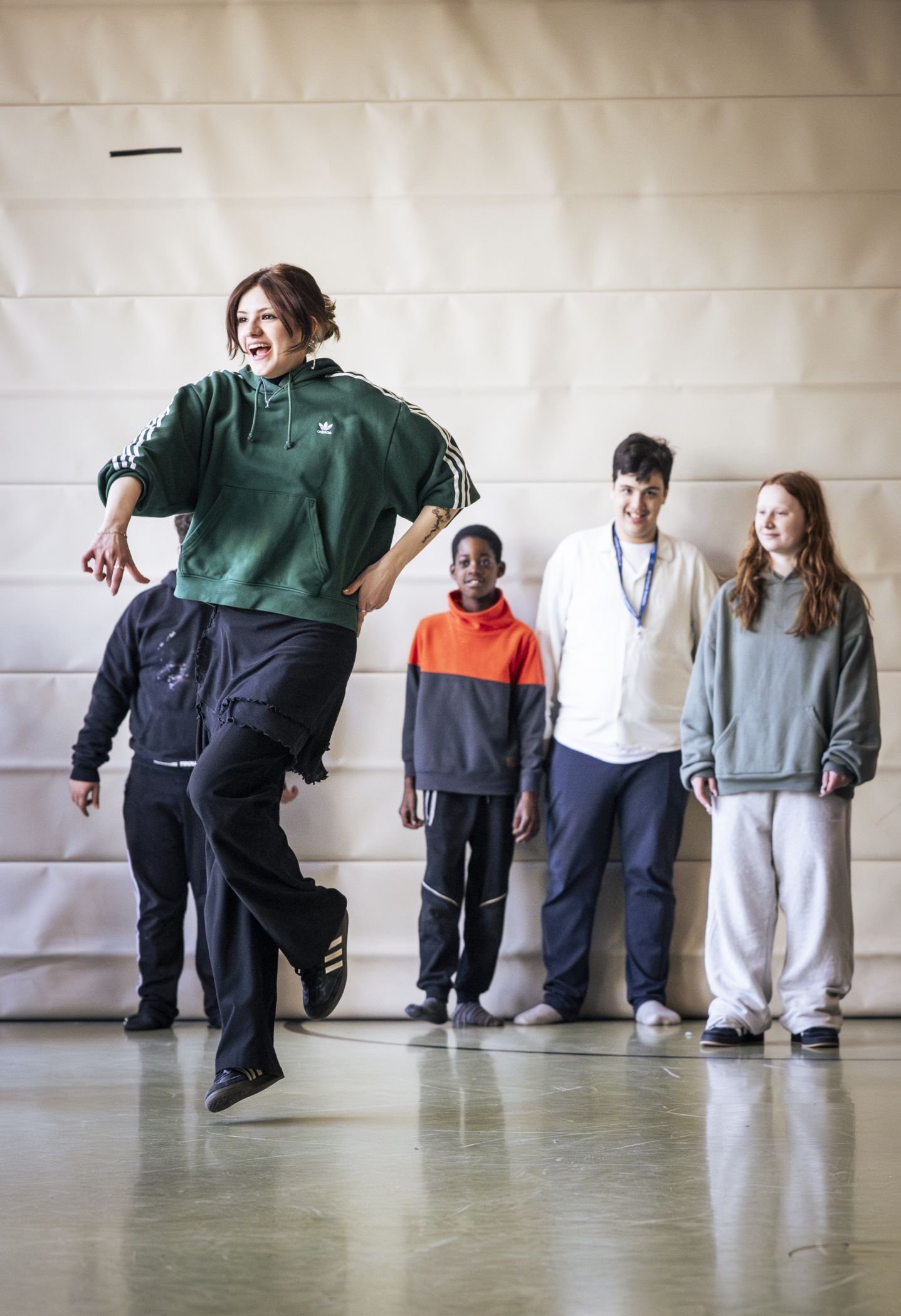 Eine junge Frau in sportlicher Kleidung führt energisch eine Hip-Hop-Tanzbewegung aus. Im Hintergrund stehen mehrere Kinder und Jugendliche und schauen ihr begeistert zu.