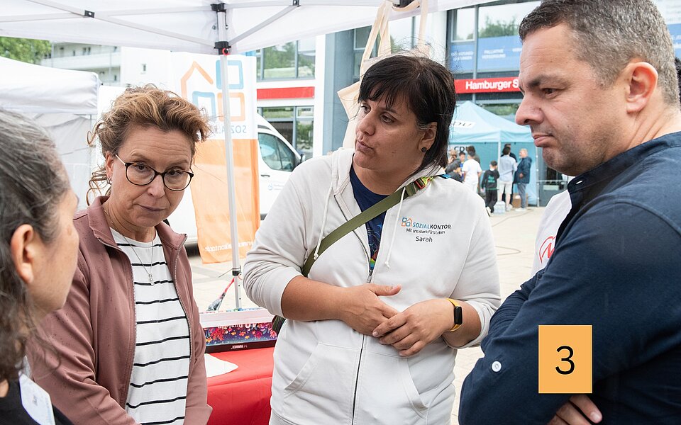 Carola Veit und Ralf Neubauer stehen beim Stadtteilfest Rothenburgsort mit Mitarbeiterinnen des Sozialkontors unter einem Pavillon und sind miteinander im Gespräch.
