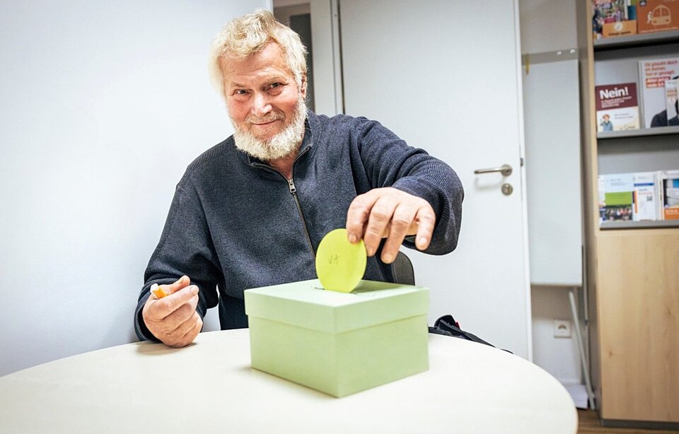 Ein älterer Mann mit weißem Bart wirft eine grüne Scheibe in den Schlitz eines hellgrünen Pappkartons. Er sitzt an einem Tisch und hält einen Stift in der linken Hand. Im Hintergrund ist ein Regal mit Broschüren und Dokumenten zu sehen.