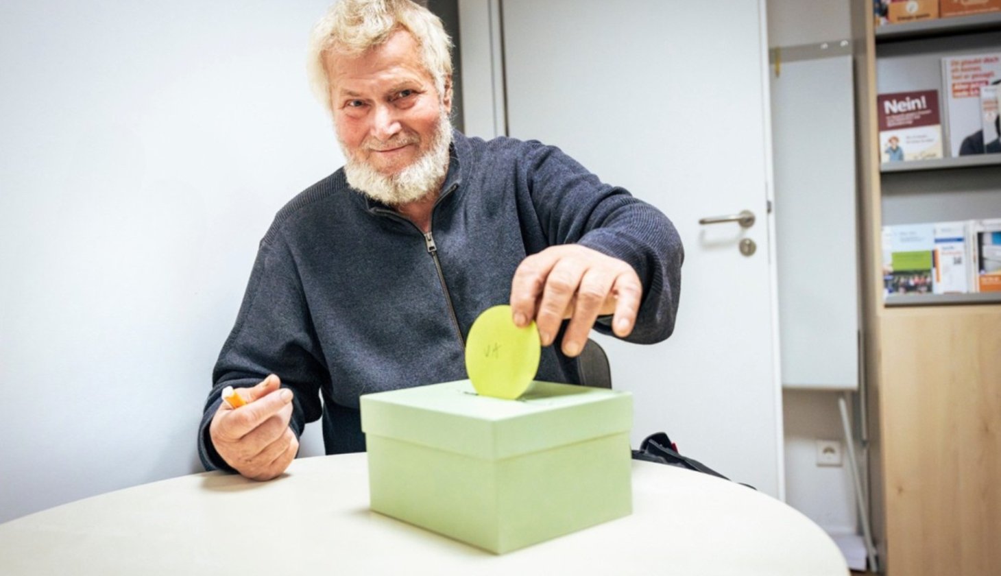 Ein älterer Mann mit weißem Bart wirft eine grüne Scheibe in den Schlitz eines hellgrünen Pappkartons. Er sitzt an einem Tisch und hält einen Stift in der linken Hand. Im Hintergrund ist ein Regal mit Broschüren und Dokumenten zu sehen.