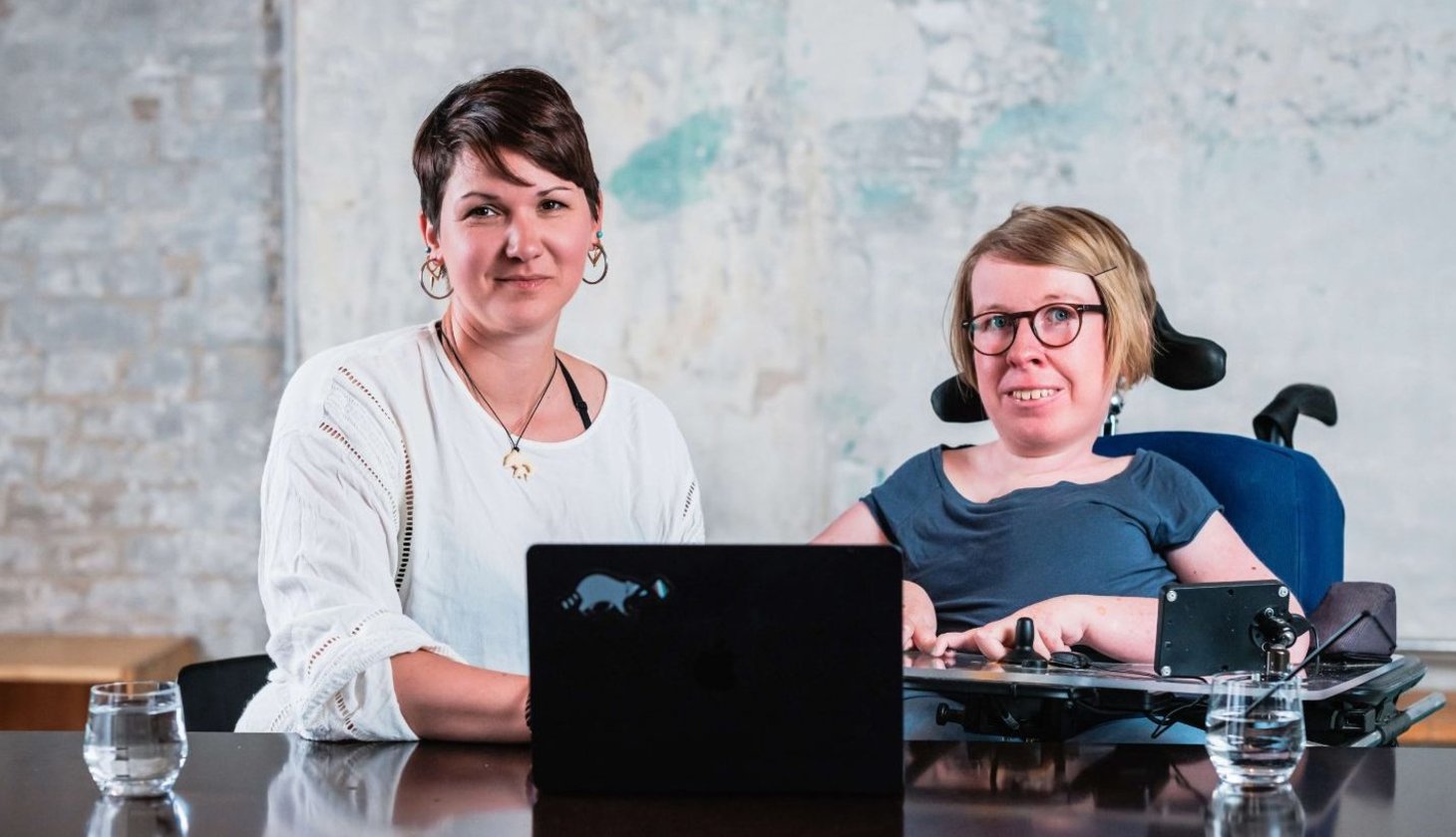 Zwei Frauen sitzen mit einem Laptop an einem Tisch, eine sitzt im Rollstuhl und beide blicken leicht lächelnd in die Kamera. Von links: Karina Sturm und Anne Gersdorff