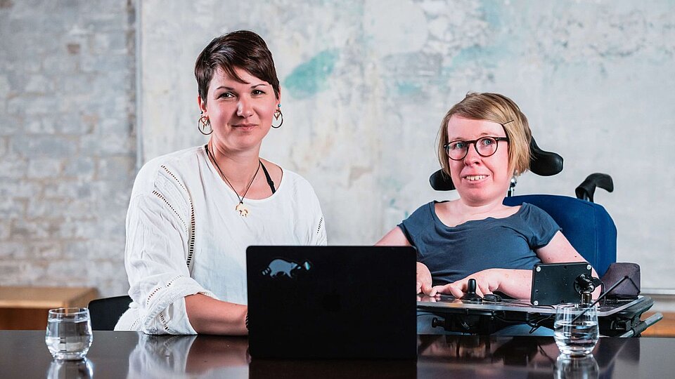 Zwei Frauen sitzen mit einem Laptop an einem Tisch, eine sitzt im Rollstuhl und beide blicken leicht lächelnd in die Kamera. Von links: Karina Sturm und Anne Gersdorff