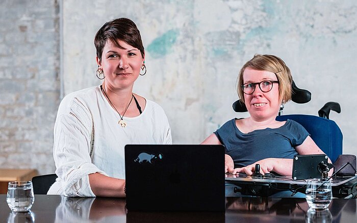 Zwei Frauen sitzen mit einem Laptop an einem Tisch, eine sitzt im Rollstuhl und beide blicken leicht lächelnd in die Kamera. Von links: Karina Sturm und Anne Gersdorff