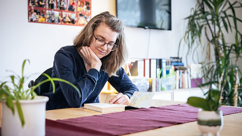 Eine Frau mit Brille sitzt an einem Holztisch in einem gemütlich eingerichteten Raum und schreibt konzentriert in ein Heft. Sie stützt den Kopf mit einer Hand ab und lächelt leicht. Auf dem Tisch stehen grüne Zimmerpflanzen und ein dunkelroter Läufer. Im Hintergrund befinden sich ein Regal mit Büchern, ein Fernseher, eine Fotowand mit Erinnerungen und weitere Pflanzen.