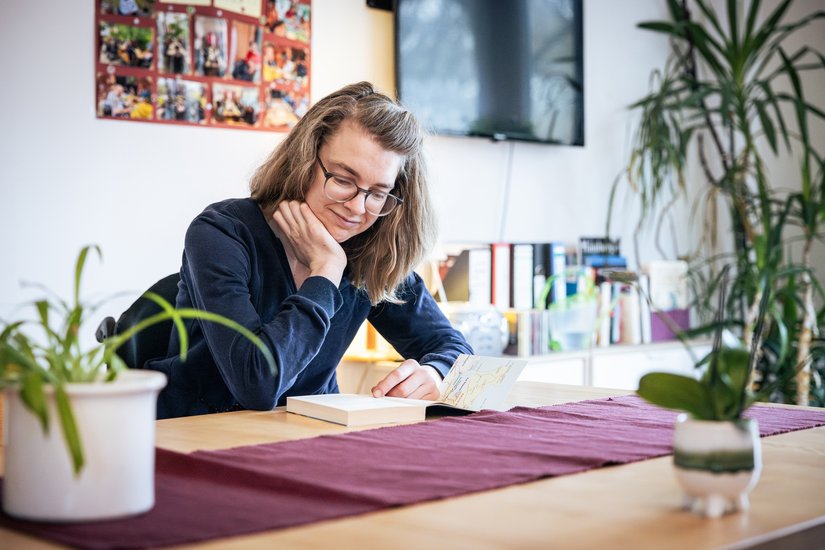 Eine Frau mit Brille sitzt an einem Holztisch in einem gemütlich eingerichteten Raum und schreibt konzentriert in ein Heft. Sie stützt den Kopf mit einer Hand ab und lächelt leicht. Auf dem Tisch stehen grüne Zimmerpflanzen und ein dunkelroter Läufer. Im Hintergrund befinden sich ein Regal mit Büchern, ein Fernseher, eine Fotowand mit Erinnerungen und weitere Pflanzen.