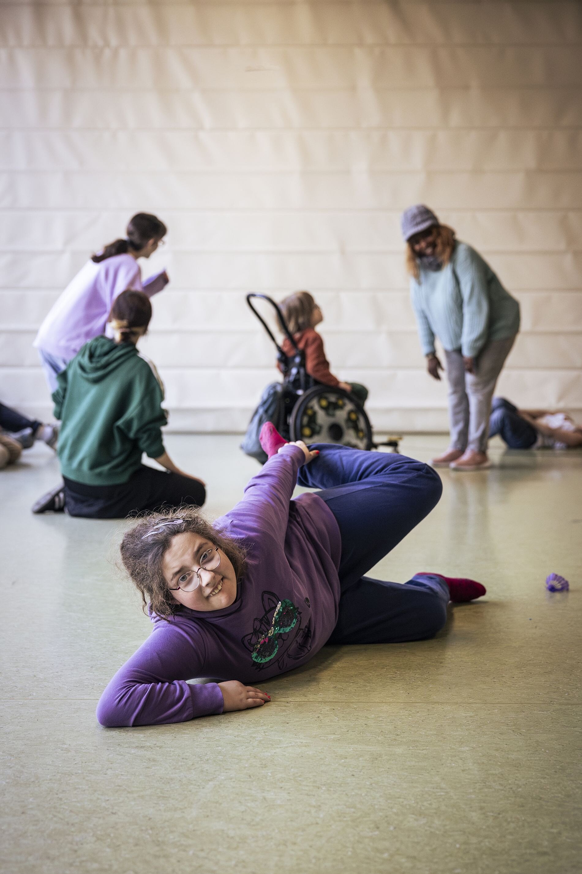 Eine Mädchen im lila Pullover liegt seitlich auf dem Boden einer Turnhalle, lächelt in die Kamera und dehnt sich. Im Hintergrund tanzen und bewegen sich weitere Kinder und Erwachsene, darunter ein Kind im Rollstuhl.
