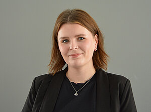 Merle Weißenberg, eine junge Frau mit schulterlangen, braunen Haaren, blickt freundlich in die Kamera. Sie trägt ein schwarzes Oberteil und ein schwarzes Jackett. Der Hintergrund ist neutral grau gehalten.