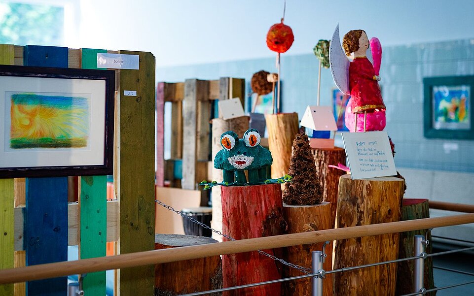 Eine Ausstellung mit verschiedenen Kunstwerken und Figuren aus unterschiedlichen Materialien. Zu sehen sind unter anderem ein gerahmtes Bild, eine grüne Froschfigur und eine rot gekleidete Engelsfigur auf Holzstämmen und Podesten.