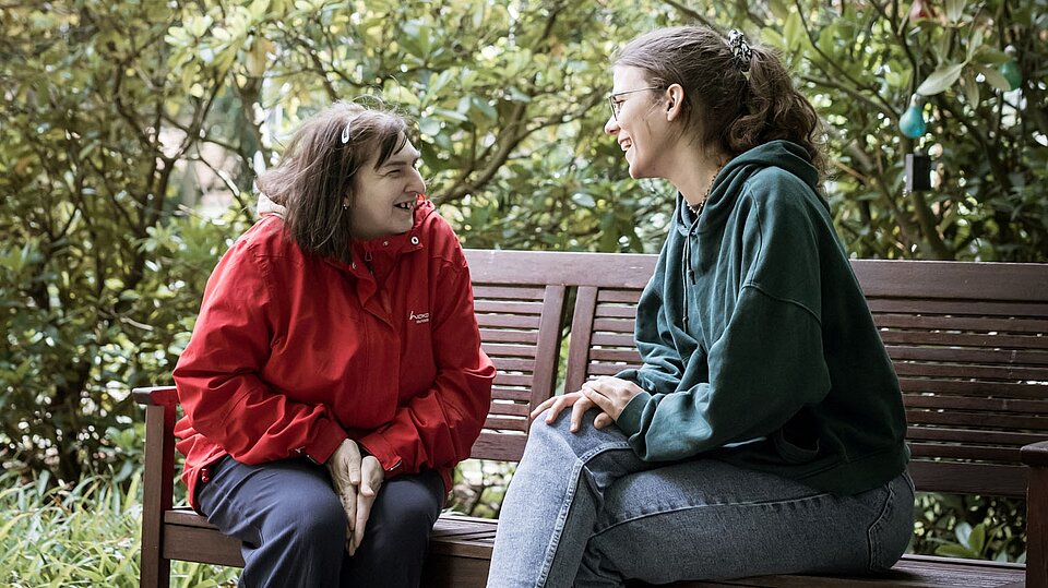 Zwei Personen sitzen sich auf einer Holzbank in einem Garten gegenüber und unterhalten sich. Eine Person trägt eine rote Jacke, die andere einen dunklen Kapuzenpullover; im Hintergrund sind Sträucher und Bäume zu sehen.