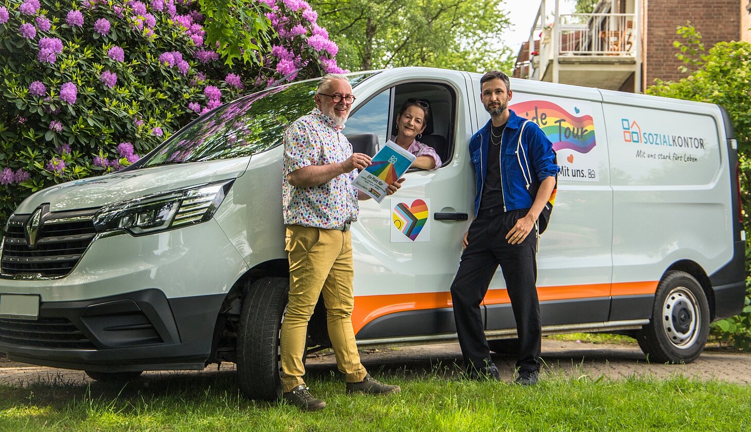 Drei Mitglieder der AG Diversity posieren vor dem Pride-Transporter des Sozialkontors. Eine Person hält Informationsmaterial zur Pride Tour in den Händen, während eine weitere aus dem geöffneten Fahrzeugfenster schaut.