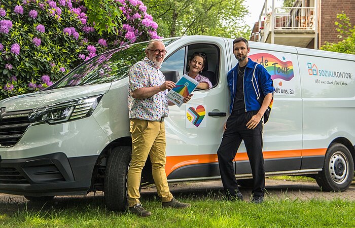Drei Mitglieder der AG Diversity posieren vor dem Pride-Transporter des Sozialkontors. Eine Person hält Informationsmaterial zur Pride Tour in den Händen, während eine weitere aus dem geöffneten Fahrzeugfenster schaut.