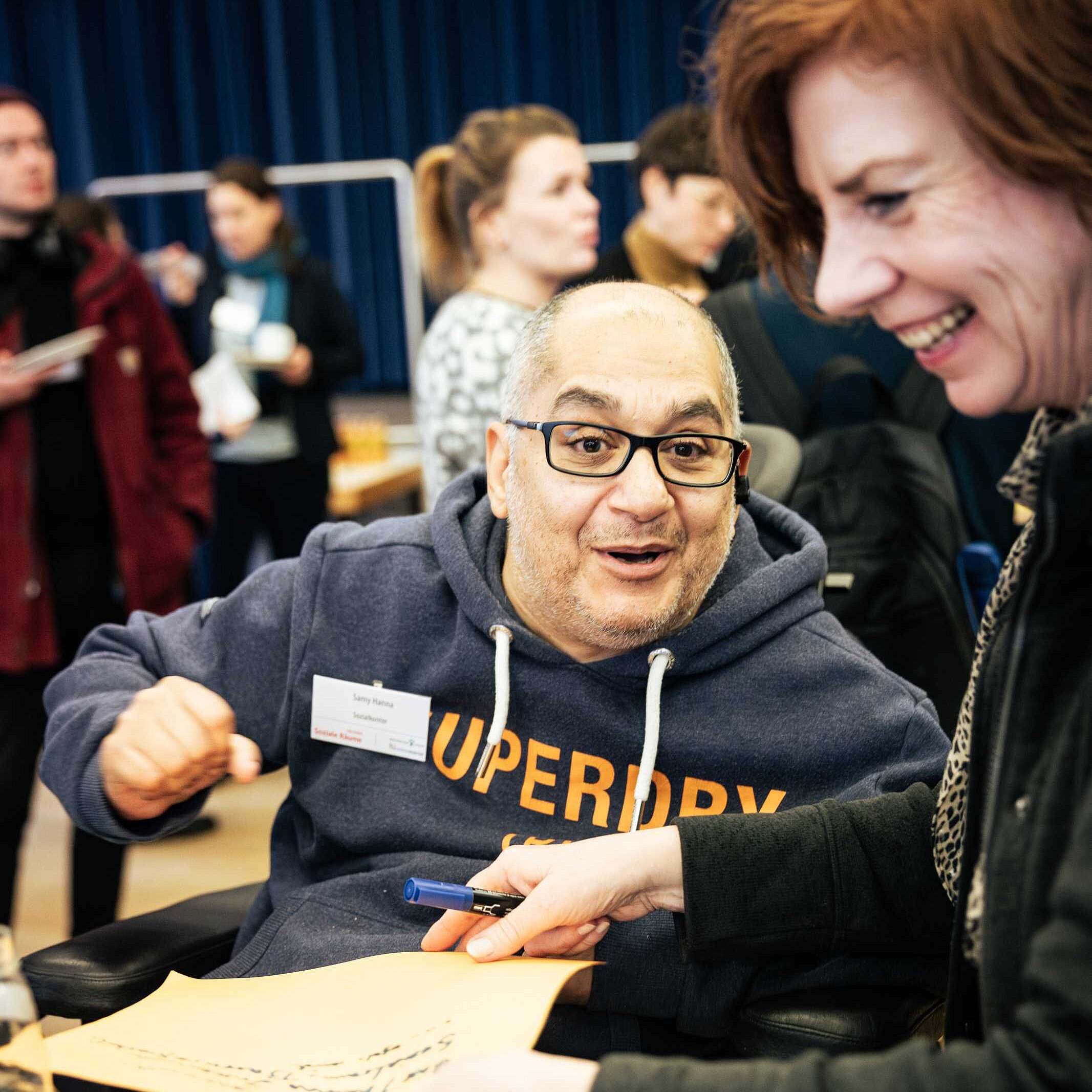 Ein Mann mit Brille und Hoodie und einem Namensschild sitzt im Rollstuhl und eine Frau mit roten Haaren steht rechts neben ihm. Sie lachen gemeinsam. Die Frau hält ein beschriebenes Blatt Papier und einen Stift in der Hand.