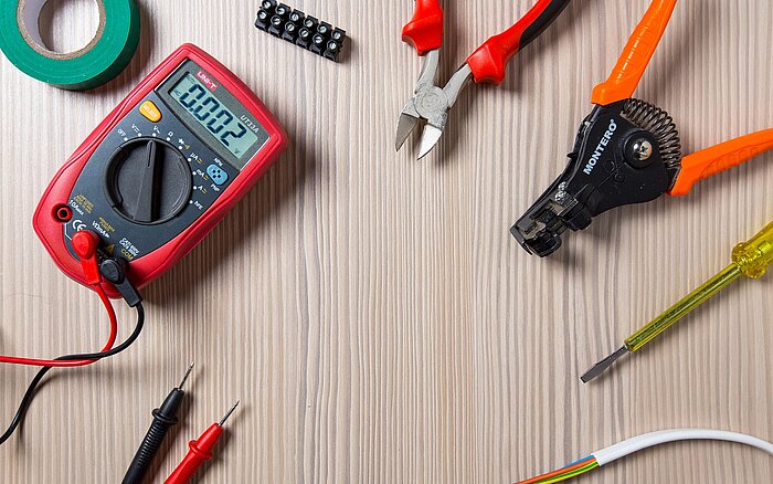 Ein Digitalmultimeter, das eine Messung anzeigt, umgeben von verschiedenen Werkzeugen, darunter eine Zange, eine Abisolierzange, ein Schraubendreher, Isolierband und Kabelverbinder liegen auf einem Arbeitstisch mit strukturierter Oberfläche.